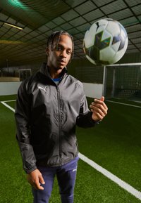 Black windbreaker jacket with a zip, featuring a white logo, worn over a blue shirt. A patterned soccer ball is suspended above.