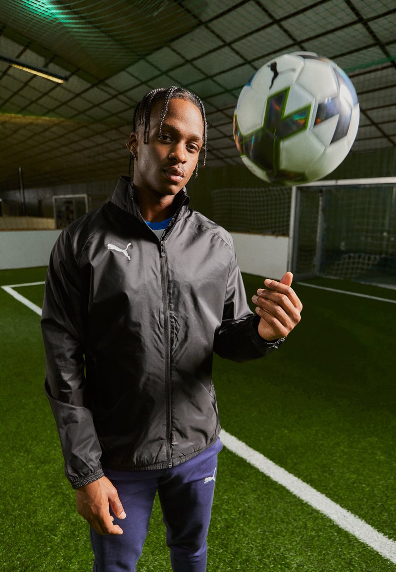 Black windbreaker jacket with a zip, featuring a white logo, worn over a blue shirt. A patterned soccer ball is suspended above.