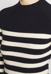 Detailní záběr černého pleteného svetru s horizontálními bílými pruhy přes hrudník a paže, s vroubkovaným jednobarevným kulatým výstřihem.