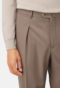 Närbild av en person som bär beige byxor med en veck, knappstängning och en ljusbeige långärmad tröja, med handen avslappnat vid sidan.