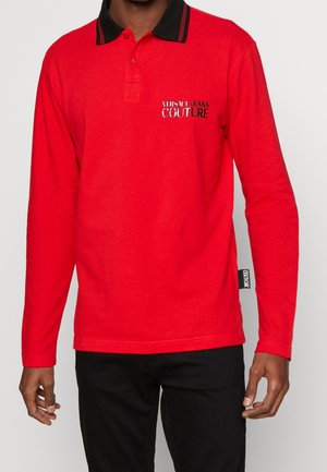 Homme portant un polo rouge à manches longues avec col noir et logo Versace Jeans Couture sur la poitrine, associé à un pantalon noir.