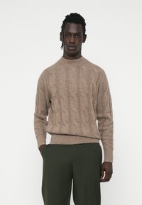 Beige Kabelstrickpullover mit hohem Kragen und gerippten Bündchen, aus weicher Wolle. Getragen mit enganliegenden dunkelgrünen Hosen.