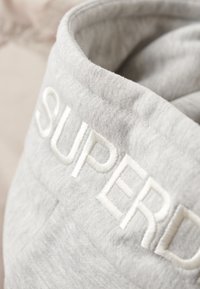 Grå stof med en blød tekstur, prydet med hvidt broderet tekst "SUPERD." Stoffet har en let strækbarhed og en glat finish.