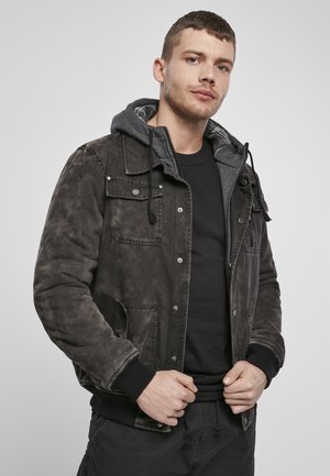 Dunkelbraune Jacke mit einem gesteppten Innenfutter, grauem Kapuzenrand, Druckknöpfen, seitlichen Taschen und gerippten Bündchen, getragen über einem schwarzen Shirt.