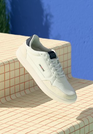 Witte leren sneaker met een textuur van grijs rubberen zool, marineblauwe accent op de hiel en veters; tentoongesteld op een gridpatroon oppervlak.