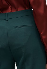 Pantalon en tissu vert avec une poche arrière cousue, associé à un chemisier lisse de couleur brun rougeâtre. Lignes épurées et coupe structurée.