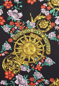 Czarna tkanina z złotym emblematem słońca, otoczona żywymi wzorami kwiatowymi w kolorze czerwonym, różowym i zielonym. Tekst na górze: "VERSACE JEANS COUTURE."
