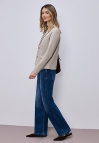 Femme marchant, portant un blazer beige, un jean bleu à jambes larges, des chaussures pointues foncées, et portant un sac à bandoulière foncé sur un fond uni.