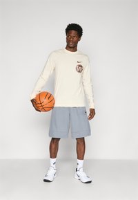 Pitkähihaista kerma-valkoista paitaa, jossa on ruskea logo, harmaat shortsit ja valkoiset tennarit, jotka pitävät oranssia koripalloa. Yksinkertainen, urheilullinen muotoilu.