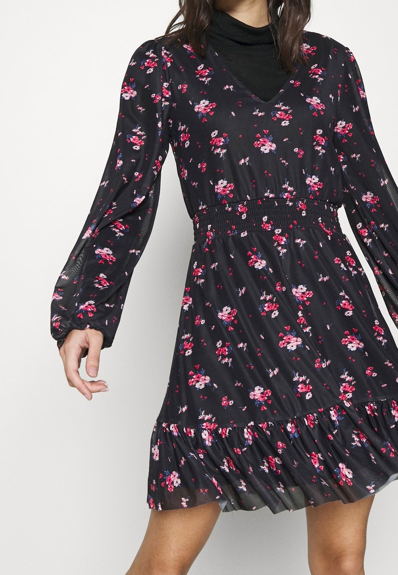 Femme portant une robe noire à fleurs à manches longues avec une taille cintrée et un ourlet volanté, superposée sur un col roulé noir.