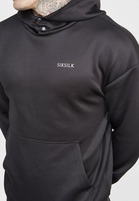 Sudadera con capucha negra de textura suave, con un bolsillo tipo canguro, botones a presión y el logo bordado de "SIKSILK" en el pecho.