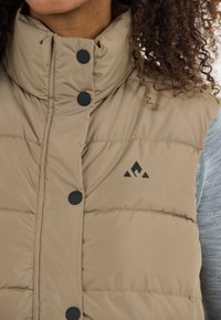 Die beigefarbene gepolsterte Weste verfügt über einen hohen Kragen, schwarze Druckknöpfe und ein Logodesign auf der Vorderseite. Der Stoff hat eine glatte, strukturierte Oberfläche.