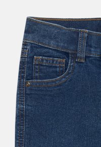 Los jeans de mezclilla azul oscuro presentan un pequeño bolsillo frontal, costuras visibles de un tono más claro y una textura de tela suave.