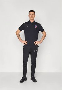 Muž stojící s rukama na bocích, oblečený v černém polu Nike Atlético Madrid a černých kalhotách Nike na jednobarevném světlém pozadí.