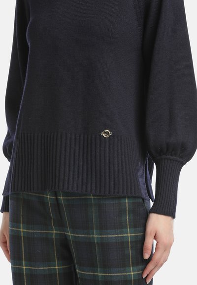 Pulover bleumarin cu tiv și manșete nervurate, având un finisaj texturat. Accesoriu din metal auriu aproape de margine. Asortat cu pantaloni în carouri.