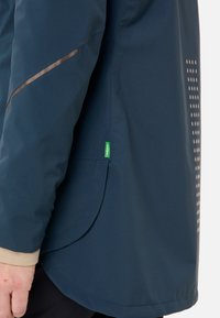 Waterdichte jas in donkerblauw met een gladde textuur, voorzien van contrasterende bruine accenten en stippendetails aan de zijkant.