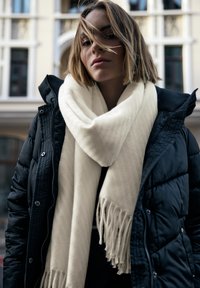 Veste matelassée noire à finition texturée, superposée sur une écharpe crème à franges, sur fond de bâtiment de couleur claire.