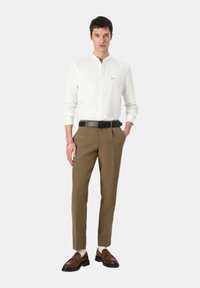 Camicia bianca a maniche lunghe con un piccolo logo, abbinata a pantaloni beige e mocassini marroni. L'outfit presenta una cintura elegante e accessori minimi.