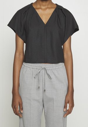 Osoba ubrana w czarny, krótki top z krótkim rękawem i plisami oraz jasnoszare spodnie z troczkiem, stojąca na tle jednolitego tła.