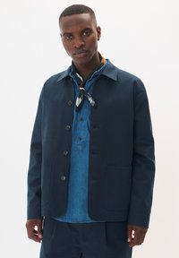 Jachetă bleumarin din material, cu închidere cu buton, buzunare aplicate și guler ascuțit, purtată peste o cămașă din denim și un fular cu model.