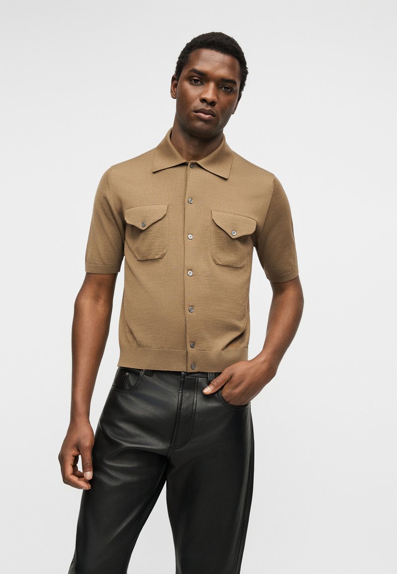 Homme portant une chemise beige à manches courtes boutonnée avec deux poches poitrine boutonnées et un pantalon en cuir noir, debout devant un fond uni.