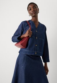 Denimjakke med dyp V-hals, lange ermer og frontknapplukking. Matchet med et denimskjørt, holder en rød skinnveske.