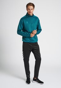 Pullover de color verde azulado con media cremallera y logo bordado, combinado con pantalones negros con franjas blancas. Ligero, con corte atlético.