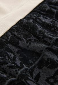 Schwarzer Samtstoff mit schimmernder Textur, der Rüschenelemente und einen glatten, beigen Taillenbund aufweist. Subtile Glitzerpunkte sind sichtbar.