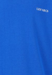 Blauwe katoenen T-shirt met een ribbelstructuur en witte tekst "LES'ARCS" op de bovenste linkerkant. Eenvoudig, klassiek ontwerp zonder extra patronen.