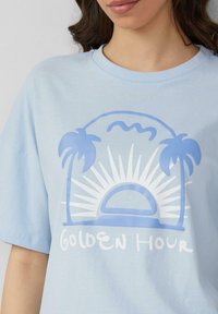 Lichtblauwe katoenen t-shirt met een witte grafische afbeelding van palmbomen, een zon en de tekst "GOLDEN HOUR" in een speelse lettertype.