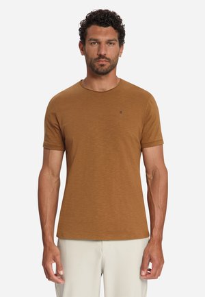 Man met kort krullend donker haar en baard draagt een effen bruine T-shirt met korte mouwen en lichtbeige broek, staand tegen een witte achtergrond.