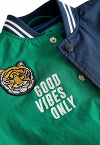 Veste verte et marine avec col rayé, écusson tigre et texte blanc « GOOD VIBES ONLY » sur le devant, boutons pression visibles.
