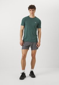 Joven de pie con una camiseta atlética verde, pantalones cortos grises, calcetines blancos y zapatillas de correr negras, contra un fondo blanco liso.