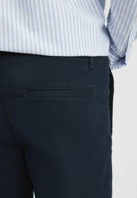 Pantalon bleu marine avec une texture lisse, doté d'une poche arrière avec des coutures discrètes et d'un logo de la marque au niveau de la taille.