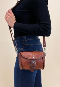 Persona che indossa jeans blu scuro e una maglietta nera a maniche lunghe, mentre tiene una borsa a tracolla in pelle marrone con fibbia e ganci metallici.