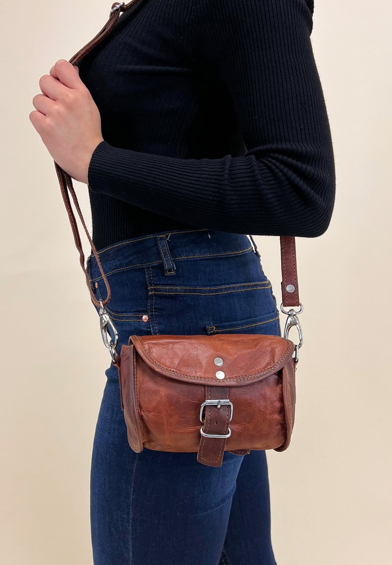 Persona che indossa jeans blu scuro e una maglietta nera a maniche lunghe, mentre tiene una borsa a tracolla in pelle marrone con fibbia e ganci metallici.