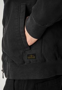 Jachetă neagră din bumbac cu fermoar frontal, mansete ribbed, buzunar lateral și etichetă de marcă pe tiv. Țesătură texturată cu un finisaj mat.