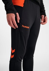 Czarne sportowe spodnie z pomarańczowym logo "hummel" i wzorem w jodełkę. Wykonane z gładkiego, elastycznego materiału, zaprojektowane z myślą o komforcie.