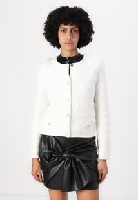 Veste blanche texturée avec un col rond et des accents de boutons dorés, associée à une jupe en cuir noire avec un grand détail de nœud.