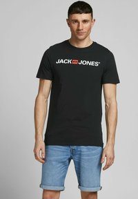 Schwarzes Baumwoll-T-Shirt mit kurzen Ärmeln, mit einem weißen Logo-Text "JACK JONES" über der Brust. Kombiniert mit hellblauen Denim-Shorts.