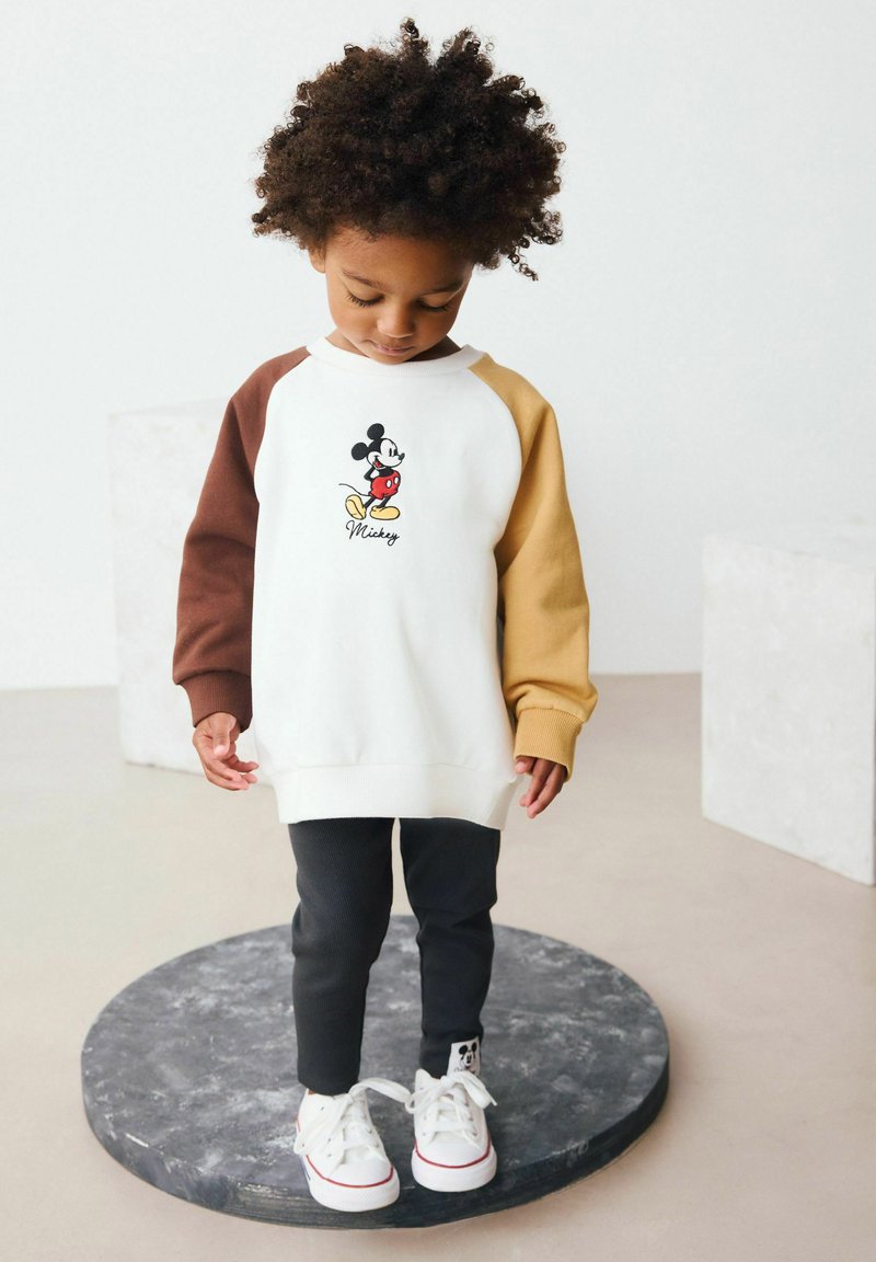 Valge dressipluus pruuni ja beeži raglanvarrukatega, millel on broderiga Mickey Mouse'i graafika. Kombineeritud mustade pükstega ja valgete spordijalatsitega.