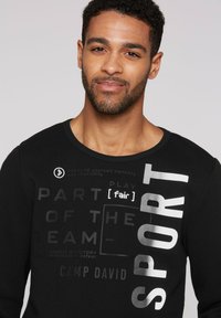 Camisa negra de manga larga con estampados gráficos texturizados en plata y gris, destacando las palabras "DEPORTE" y "JUGAR [justo]" con un escote de cuello redondo.