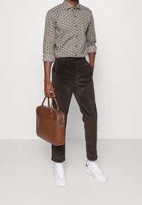 Bruine corduroy broek, overhemd met patroon en knopen, witte sneakers, bruine leren tas met handvatten en zilveren rits.