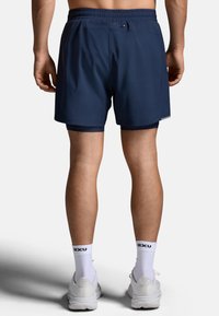 Granatowe sportowe szorty z elastycznym pasem, tylną kieszenią i siateczkową podszewką. Noszone z białymi skarpetkami z logo i butami.