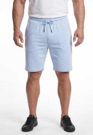 Homme portant un short bleu clair à cordon, une chemise blanche et des baskets noires, debout les bras détendus.