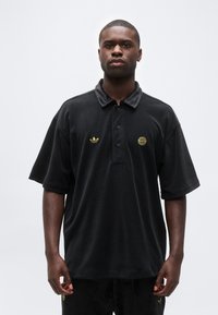 Άνδρας που φοράει μαύρο πόλο με κοντά μανίκια, με χρυσά λογότυπα Adidas και FC Bayern, στέκεται μπροστά σε απλό φωτεινό φόντο.