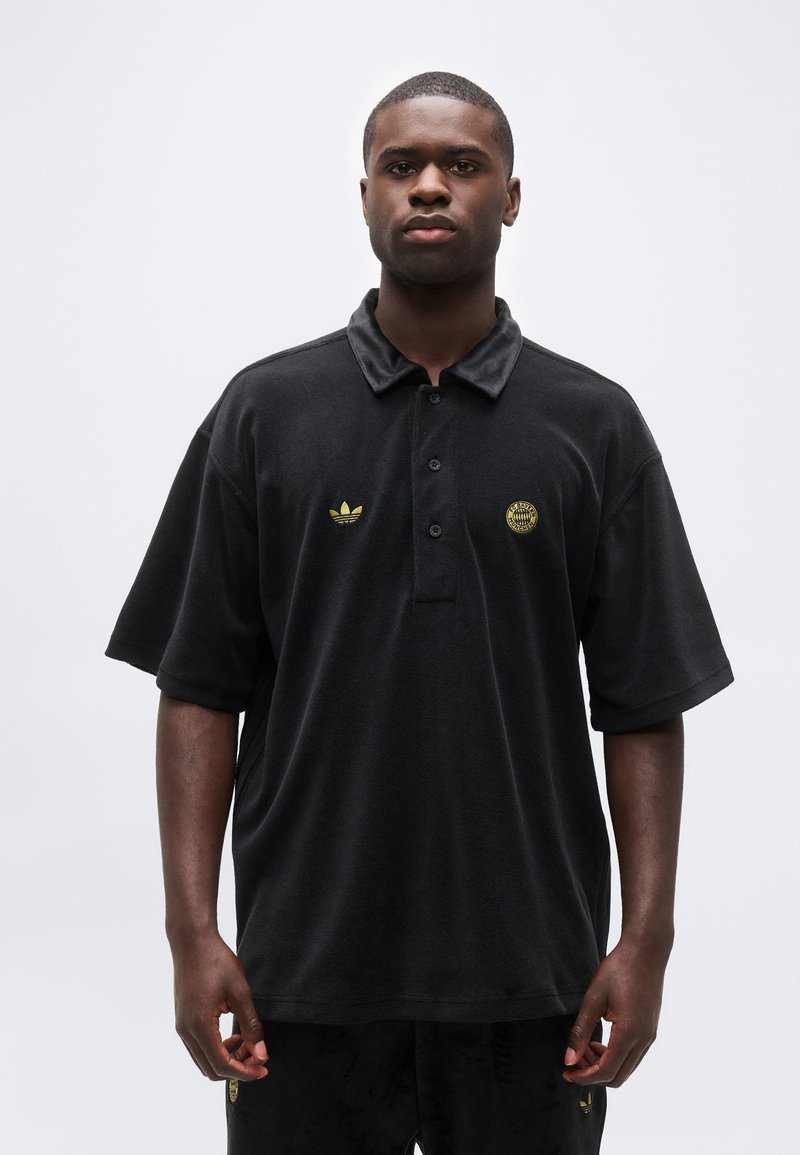 Άνδρας που φοράει μαύρο πόλο με κοντά μανίκια, με χρυσά λογότυπα Adidas και FC Bayern, στέκεται μπροστά σε απλό φωτεινό φόντο.