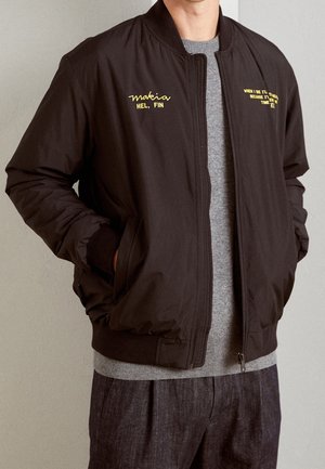 Homme portant une veste noire zippée avec un texte jaune sur la poitrine, un pull gris, un pantalon foncé, les mains dans les poches de la veste, debout à l'intérieur.