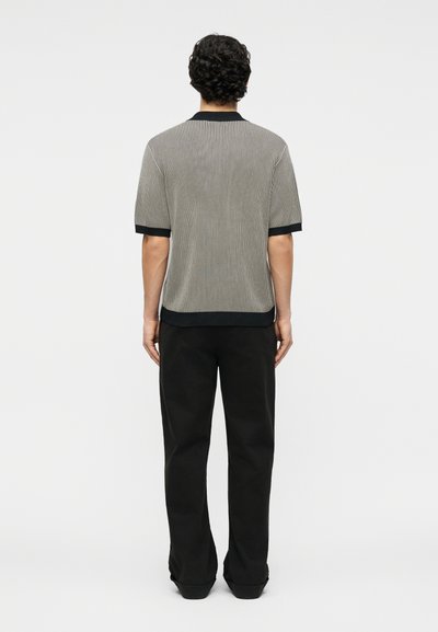 Homme aux cheveux bouclés et foncés, vu de dos, portant une chemise à manches courtes rayée verticalement avec des bordures noires et un pantalon noir.