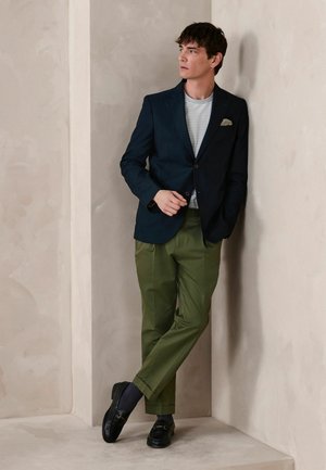 Man wearing navy blazer, striped shirt, olive trousers, and black loafers leaning against a beige wall, looking to the side.